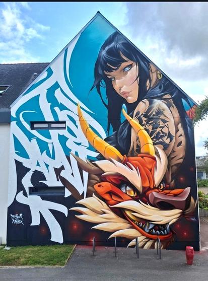Streetartwall. A beautiful mural of a young woman with a manga-style dragon was sprayed/painted on the exterior wall of a one-story modern building (city hall). She has long black hair, blue eyes and an upper arm tattooed with tribals. In front of her is a fierce-looking red dragon with yellow horns and a beige mane. The young woman looks at the viewer with a fierce gaze. The background consists of white graffiti lettering on a turquoise background.
Info: A personal and contemporary version of Mulan by the two artists Mr. Manz and Asem Navarro, which is intended to stand for the courage of women and their fight for equal rights as well as for the modern way of graffiti in the world of art.