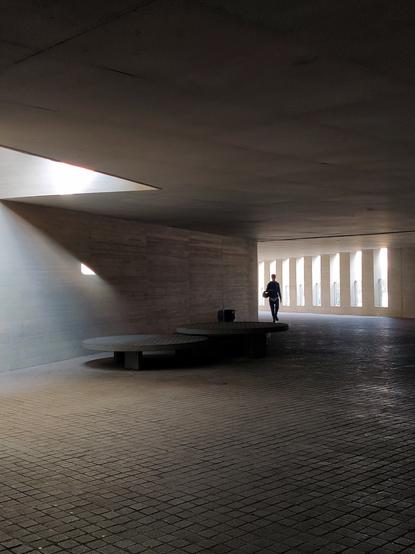 Corredor exterior de la facultad de Medicina. Se ven unas mesas de hormigón sobre las que cae la luz. Al fondo hay una fila de ventanas. Por delante de ellas pasa una persona.