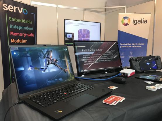 Table showcasing some of the work done by Igalia. On top of it, an ARM laptop running Portal 2, a display showcasing Servo's features, a Raspberry Pi in a case, a Steam Deck and some stickers. In the background, a Servo browser banner, a TV showcasing our work on Webkit and an Igalia banner.
