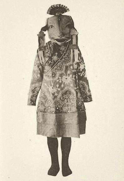 Mujer con traje tradicional con mangas largas y estrechas y falda por las rodillas. Tiene una máscara con un abanico en la cabeza y cintas en las orejas, con la boca abierta.
