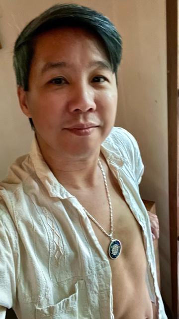 A person with short, dark hair and a slight smile is wearing an unbuttoned white shirt, revealing a necklace and part of their chest. The background features a neutral-colored wall.
