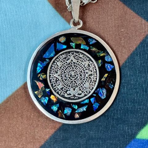 A circular pendant featuring a central silver medallion with intricate designs, surrounded by small, colorful, iridescent fragments set against a black background. The pendant is attached to a silver chain, resting on a textured surface with various colors.