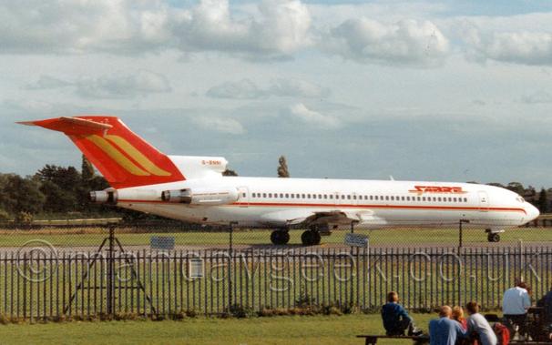 Side view of a white, three-engined jet airliner with the engines mounted on the rear fuselage, taxiing from left to right.
There is a red and gold stripe running along the body, with a red tail with a pair of gold stripes running diagonally from the top.
There are red "Sabre" titles on the upper forward fuselage with golden drop shadows, and the registration "G-BNNI" on the centre engine intake in red.
Green grass fills the foreground, with a group of people sitting around and on some wooden picnic benches in the lower right corner.
A black metal fence stands between them and the plane.
Trees line the skyline, under layered grey clouds.