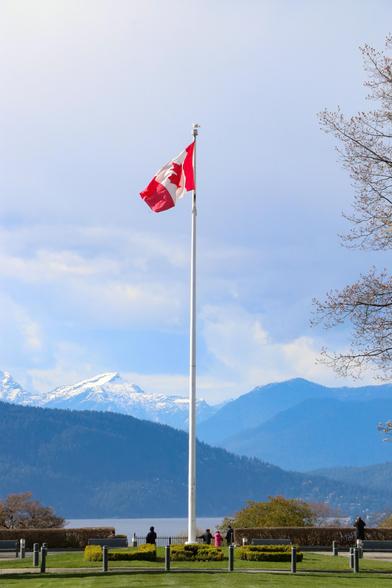 A Canada flag ripples in a gentle breeze on its flagpole at the University of British Columbia. Mountains and water are seen in the background, along with a mostly-cloudy sky, typical of the Vancouver area.