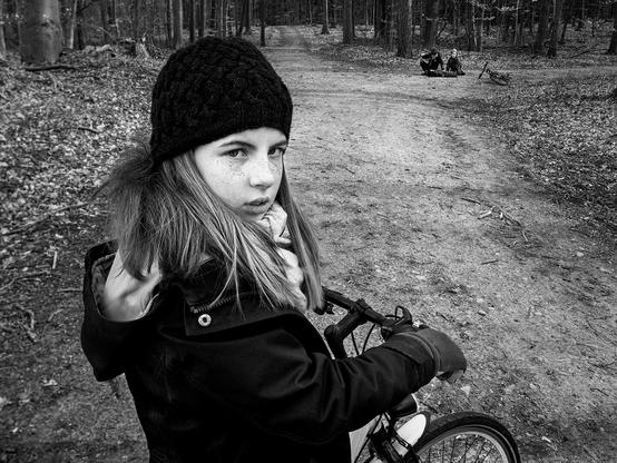 Junges Mädchen ist mit einem Fahrrad auf einem Waldweg. Im Hintergrund schlagen zwei Jungs an einer Weggabelung einen weiteren Jungen zusammen.