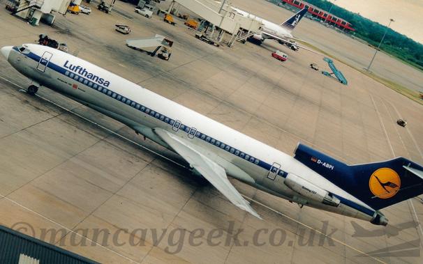 Side view of a white, 3 engined jet airliner with the engines mounted on the rear fuselage, taxiing from right to left, taken at a slight angle.
The plane has a silver belly, and a blue stripe running along the body, covering the passenger cabin windows.
There are blue "Lufthansa" titles on the upper forward fuselage.
The tail and centre engine intake are dark blue, with a yellow circle containing a stylised blue flying bird on the tail, and the registration "D-ABPI" in white on the centre intake.