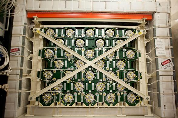 The HALO Detector. We see green-painted lead bricks, encirclind white plastic cylinders each holding 4 neutron detectors made using Helium-3. A steel frame supports the detector. It is surrounded by water boxes to protect it from ambient neutron radiation, naturally emitted by the rock in the walls of the laboratory.