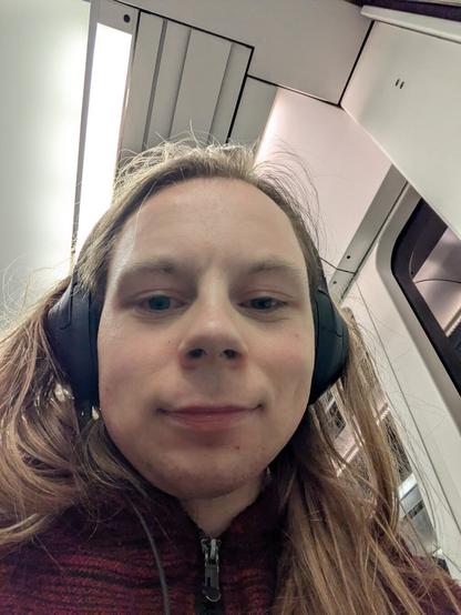 The image captures a young man with long hair wearing headphones, a solo traveler on a train.

The person is positioned within the confines of the train car, indicated by the ceiling panels and the window visible in the background.

The lighting is bright and artificial.