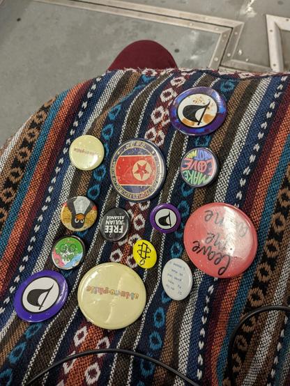 This image depicts a bag overflowing with a diverse assortment of buttons and pins in various vibrant colors against a backdrop of a grey floor on a public train.