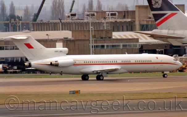 Side view of a white 3 engined jet airliner with the engines mounted on the rear fuselage, taxiing from left to right.
There is a gold and red stripe running along the body just below the passenger cabin windows, with a second gold stripe just above the windows, and a reversed Bahrain flag (Large red field next to a large white field, separated by a jagged edge - normally displayed the other way around) on the tail.
The registration "A9C-BA"is on the centre engine inlet in black.
A black runway stretches across the bottom of the frame, separated from this plane by a strip of grass.
A brown building stretches across the background, with the grey rear fuselage and tail of a very large jet airliner is on the right of the frame.
Leafless trees stand out against grey sky beyond the building.
