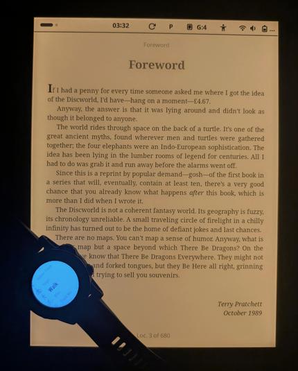 PineNote in a dark room with its warm front light which is a gentle glow. There's a Garmin watch on top of the screen blasting out harsh blue light. The PineNote is open to the Foreword of Terry Pratchett's The Color of Magic.