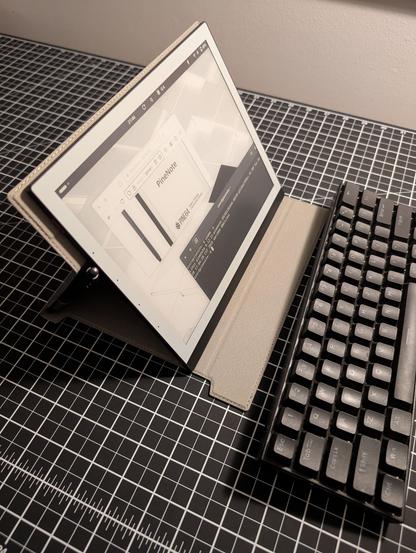 The PineNote is shown from a sideview resting on a table with black cutting mat. The folio is folded to prop the tablet up like a laptop screen and a wireless keyboard is in front of the screen. The screen is showing a small browser window and a terminal window with uname -a output
