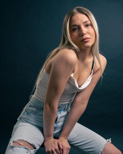 The image features a person with long blonde hair, wearing a white top and ripped blue jeans, posing against a dark background.
