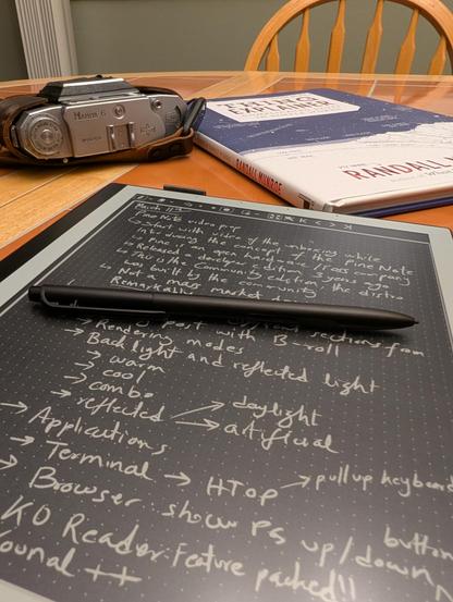 PineNote on a table with a book and camera in the background. PineNote is in black and white inverted mode (white writing on background) and there are hand scribbled outline of things to cover in the video (broadly things in previous posts). Text is crisp and easy to read despite subpar handwriting.