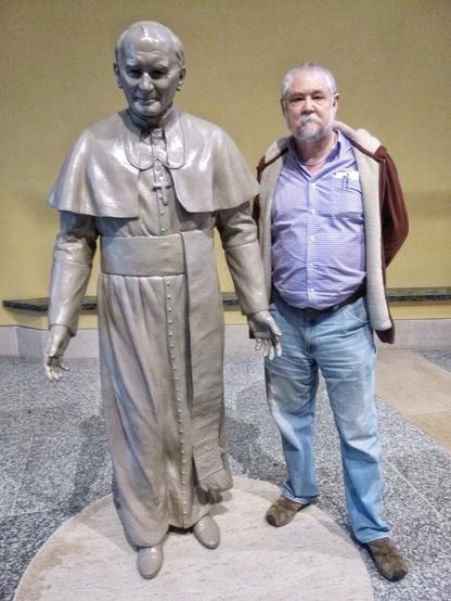 Man with white beard, dark red jacket, and jeans poses with a life-sie statue of Pope John Paul II.