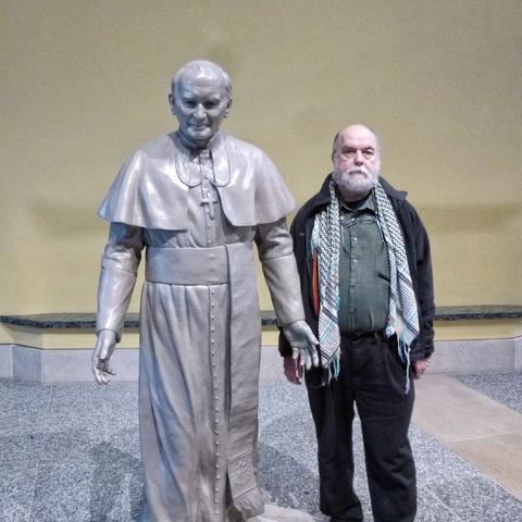 Man with white beard, dark jacket and jeans poses with a life-size statue of Pope John Paul II.