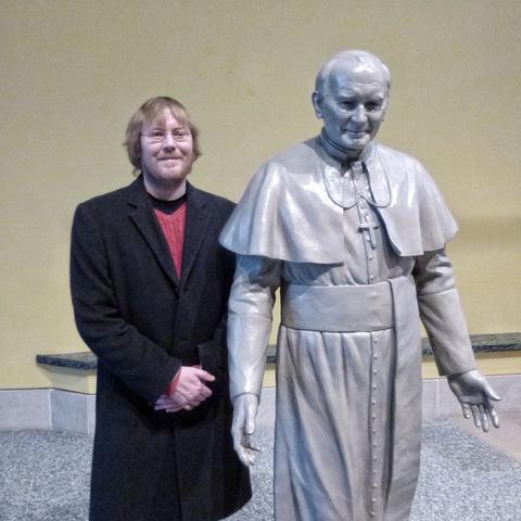 Man with red beard and long, black coat poses with a life-size statue of Pope John Paul II.