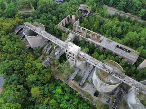 Zdjęcie lotnicze opuszczonego kompleksu przemysłowego, porośniętego bujną roślinnością. Widoczne są zrujnowane betonowe budynki, duże cylindryczne zbiorniki i korytarze technologiczne, które uległy degradacji. Natura stopniowo przejmuje teren, wypełniając puste przestrzenie gęstymi drzewami i krzewami.