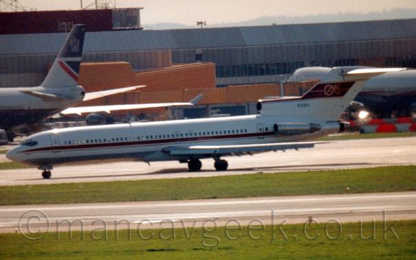 Slightly soft focus photo of a 3 engined jet airliner taxiing from right to left.
The plane's engines are mounted on the sides and top of the rear fuselage.
There is a red stripe running along the body, and another stripe running along the top of the centre engine and tail, overlaid with a gold "GM" logo.
Green grass either side of a runway fills the foreground, with a large grey terminal building with yellow airbridges connected to very large dark blue and grey jet airliners stretches across the background, with bright but hazy sky visible right at the top.
