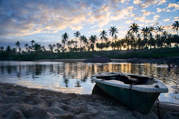 A sunset at a beach, with the sun setting on the land side. Up-front the beach with an empty boat are visible, then a river which separates the beach from the land. On land palm trees and a hut are backlighted by the setting sun on the horizon. On the upper part of the photo the blue sky and some clouds are seen.