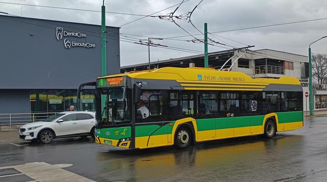 Trolejbus TLT Tychy (dominujące kolory: żółty i zielony) w deszczowej scenerii przed tyskim dworcem kolejowyn.