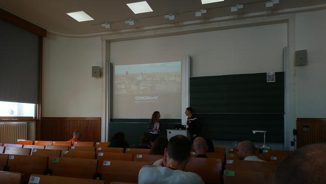 In einem Hörsaal sitzen Zuhörer mit dem Rücken zur Kamera. Der Sprecher und Moderator stehen seitlich vor der Beamerleinwand und der Tafel. Die Präsentation zeigt den Schriftzug “osm2world"