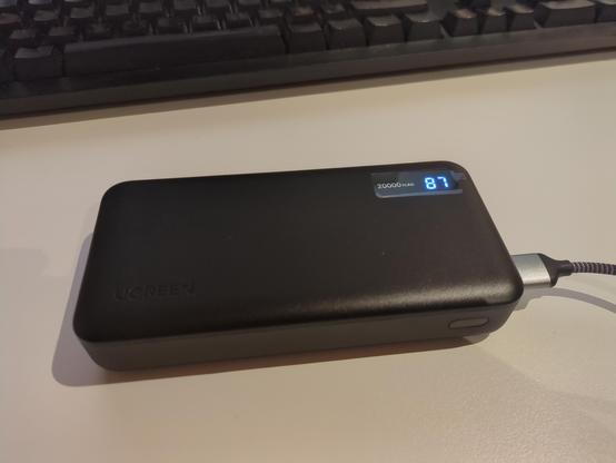 A black power bank on a white desk. It has a small display in the top right corner which shows a charge of 87%.