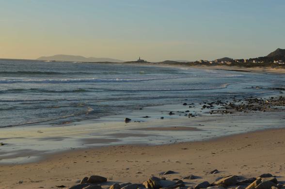 Imaxe dunha praia. Ao lado esquedo está a area e ao dereito o mar. Ao fondo hai un faro e algunhas vivendas. Na parte inferior vense algúns pedras que están situadas na area. Hai ondas pequeniñas. É día.