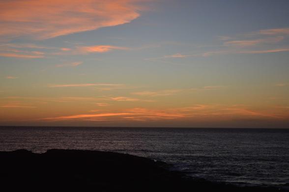 Imaxe do solpor co ceo laranxa sobre o orizonte do mar. O sol xa non está presente. Na parte inferior vense unhas rochas (xa escuras pola falta de luz). Hal algunhas nubes altas que están de cor laranxa.