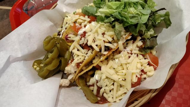 Three tacos in a paper lined basket. The shredded cheese obscures the ingredients beneath. There is some lettuce to one side of the tacos, and some jalapeños to the other.