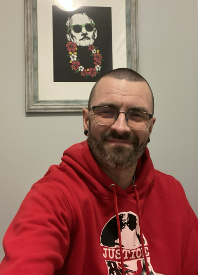 Man smiling at the camera. Wearing a red hooded sweatshirt with the text “Justice” and the design of an Indigenous woman with red hand print across her mouth.