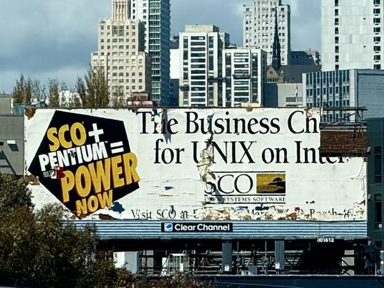 A weathered and partially torn billboard in front of a city skyline promotes SCO UNIX software on Intel hardware. The left side reads “SCO + Pentium = POWER NOW” in bold, yellow and white letters on a black geometric background. The right side, more deteriorated, reads “The Business Choice for UNIX on Intel” with the SCO Systems Software logo below. The ad shows signs of age, with peeling paint and faded text. The billboard is mounted on a structure labeled “Clear Channel.” Tall buildings and trees are visible in the background.