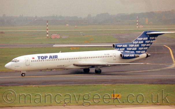 Side view of a white, 3 engined jet airliner with the engines mounted on the rear fuselage, taxiing from right to left on a black taxiway.
The plane has blue "Top Air" titles on the upper forward fuselage, with the registration "TC-IYA" on the upper rear.
The tail is dark blue, with small, repeated white "Top Air" titles.
The engine pods are the same blue, with the same "top Air" titles.
Green grass fills the foreground, with more grass and black runways in the background, with trees vanishing into grey haze in the distance.