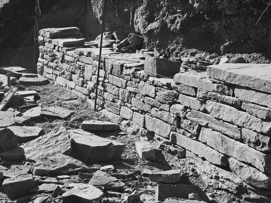 A black and white photograph of a drystone wall, partially built, in the bright sun.