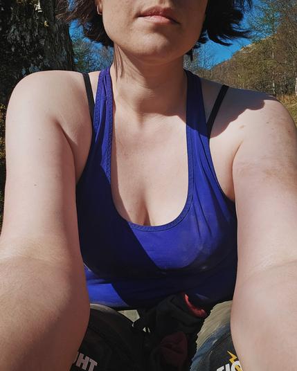 A woman sitting outdoors in sunlight, wearing a blue tank top and work trousers. Her face is partially out of frame, capturing her chin and mouth, with blue sky and trees behind her.