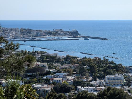 The bay of Foro with a passenger ferry leaving. The sky is light blue and clear, while the water is dark blue. There are many buildings visible, but too small to make out individual details.