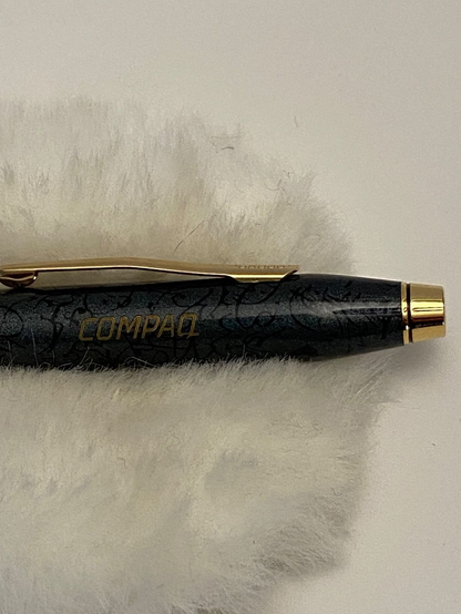Close-up of a dark, patterned fountain pen with gold accents, resting on a soft white surface. The word 'COMPAQ' is engraved in gold lettering on the pen's cap. The pen clip and trim are also gold-coloured, and the design suggests it is a promotional item, produced by Cross.