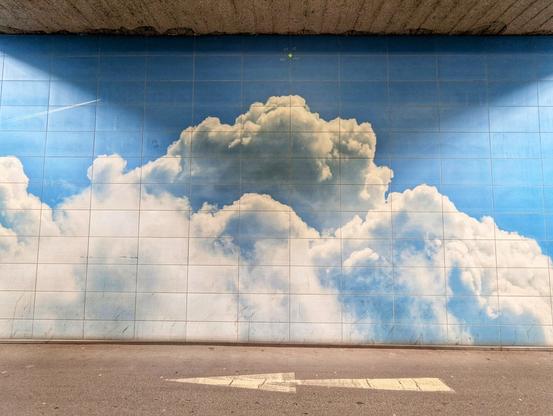 A low-angle shot shows a section of wall under an overpass. The upper portion of the wall is tiled and painted with a bright blue sky and fluffy white clouds. A faint white contrail from an aircraft stretches across the upper left of the painted sky. Below the painted section, on the ground, is grey asphalt with a large, faded white arrow pointing to the left. The concrete ceiling of the overpass is visible at the very top of the frame.