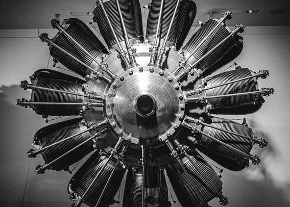 Black and white image of a vintage radial aircraft engine, viewed head-on, with multiple cylindrical pistons arranged in a circular formation around a central hub. The metal parts are highly detailed, showcasing bolts, fins, and rods with strong industrial textures.