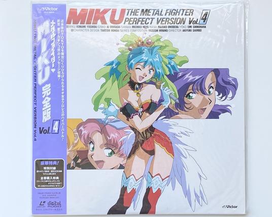 A photo of the cover for the anime Metal Fighter Miku.
It's the big Laserdisc version.
The cover shows Miku, the young anime girl with light skin and cyan colored hair.  She's wearing a read showgirl dress with white feathers.