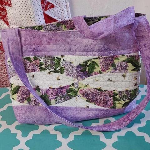 a photo of the tote bag, it is lavender colored with handles on it, there is a wide white strip across the middle with curved arcs of green and lavender foliage on them, 4 of them, converging