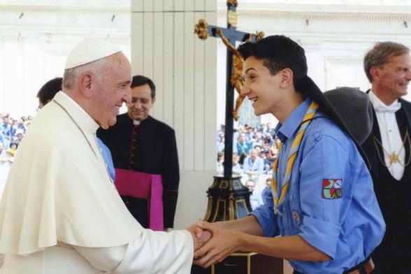 A photo of me and Papa Francesco talking. He is on the left, smiling and shaking my hand. I am on the right, in scout uniform, with a cheerful expression.