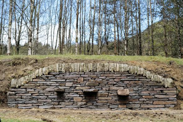 A head-on shot of the finished drystone wall, showing the full arch and the even, tight stonework. The three large protruding stones, meant to hold a bench, are clearly visible. Behind, a stand of thin trees in early leaf frames the scene.