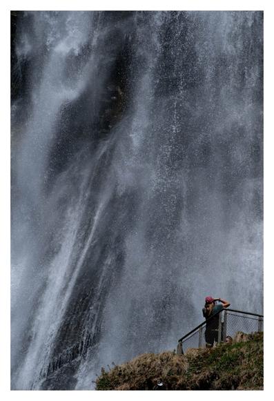 Foto im Hochformat. Teleaufnahme, der Großteil des Bildes sind die Wassermassen eines Wasserfalls, die herab fallen. Weiße Gischt, dahinter dunkle Felsen. Rechts unten ist eine Plattform auf einem begrasten Hügel angeschnitten im Bild. Darauf steht eine Person und fotografiert den Wasserfall. Auch wenn man den ganzen Wasserfall nicht sieht, durch die Größe bzw. Winzigkeit der Person, wird deutlich, dass dieser sehr groß sein muss.