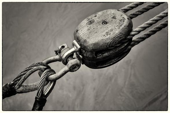A conference connection economy: A black and white photograph of a wooden pulley. Two sisal ropes loop around the pulley on its right. On the left of the pulley is a metal shackle with two metal ropes attached to it.