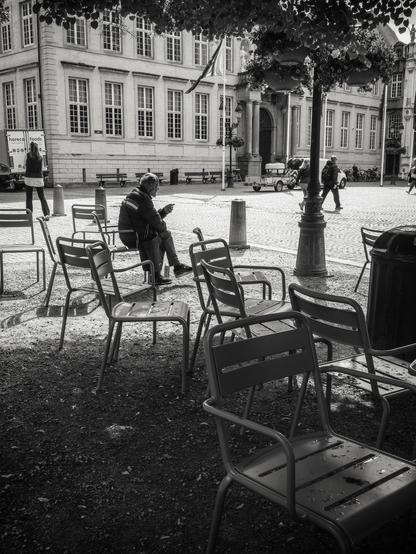 The image is a black and white photograph of an outdoor scene in a public square in Bruges, Belgium. In the foreground, several metal chairs are arranged on a gravel surface, with one person sitting on one of the chairs. The chairs are empty except for this individual. 
In the background, a large building with classical architecture, featuring numerous windows and a prominent entrance, is visible. A flagpole with a flag is also present. People are walking in the square, and a food delivery truck is parked nearby. The cobblestone pavement adds to the historical ambiance of the scene.