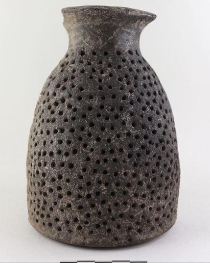 Photo: M. Simon/ Novetus

The picture shows a pottery vessel: a dark grey, round-bodied, ceramic jar with numerous small holes distributed across its surface. The holes create a patterned effect.