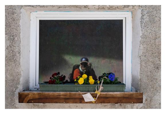 Foto im Querformat. Ein rechteckiges Fenster in einer hellen verputzten Wand. Vor dem Fenster ist eine braune Fensterbank. Darauf steht ein länglicher Blumentopf mit drei verschiedenfarbigen Blumen. Weinrot, gelb und blau von links nach rechts. Außerdem hängt noch ein kleiner Flugdrache an der Fensterbank. Im Fenster spiegel ich mich, orangefarbenes Ahirt, Rucksack, Basballkappe, leicht gebeugt, die Kamera vorm Fressbrett. Der Fokus ist aber auf den Blumen, ich bin leicht unscharf.