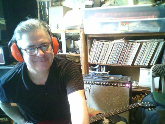 A happy old dude in a black crew shirt and glasses, wearing orange safety headphones, with a Fender Super Reverb 4x10 Amplifier behind him, with the  red jewel light glowing.