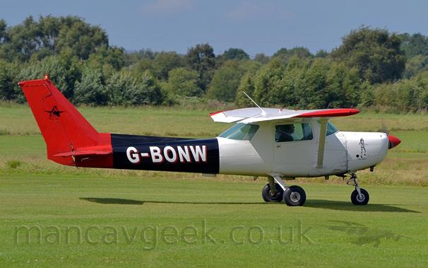 Side view of a high-winged, single-engined light aircraft taxiing from left to right on grass.
The plane has a white forward fuselage, a black rear fuselage, and a red tail, with the registration "G-BONW" on the rear fuselage in white, with a red drop shadow.
The spinner on the propellor is also red.
There is a black and white drawing of a female figure dressed in a stereo-typical striped prison outfit with a large, spherical weight attached to her ankle, above black text reading "The Jailbird" on the engine cowling.
A couple of people can be seen through the window in the cockpit door.
Mown green grass fills the foreground, with un-mown grass in the background leading up to trees in the distance, under grey-blue sky.